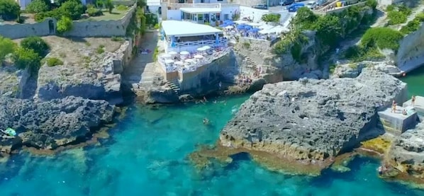 A coastal area with rocky terrain and clear blue waters. There is a building with a terrace overlooking the water, and a few people are visible on the rocks.