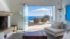 Un soggiorno moderno con vista sul mare, una porta vetrata che dà su un’area salotto esterna e un divano bianco.