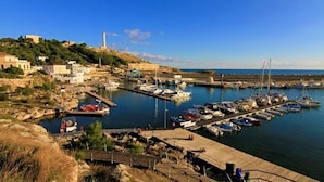 Una marina costiera con imbarcazioni ormeggiate, un faro e edifici lungo la riva.