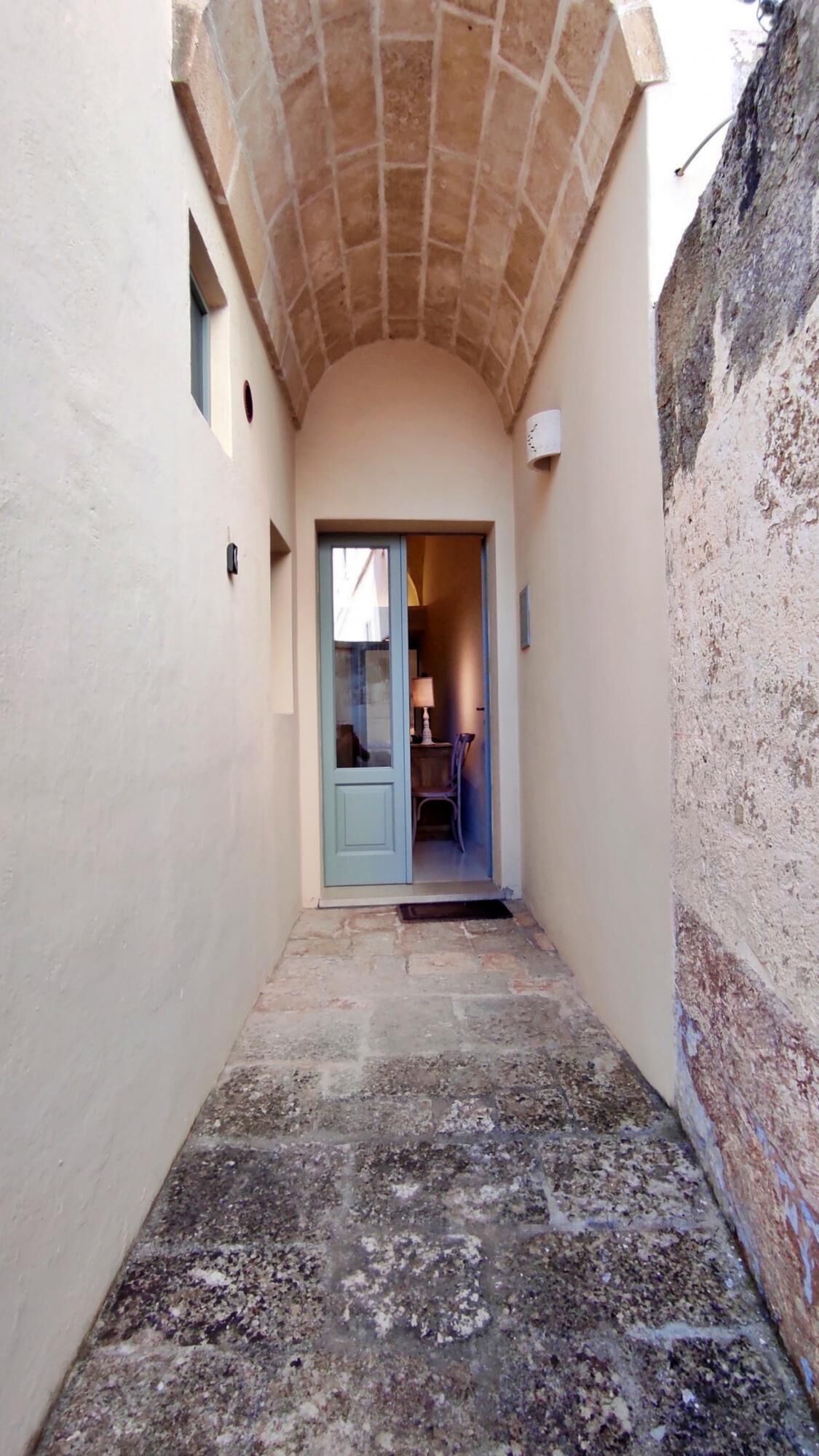 A narrow corridor with a tiled floor, a door leading to a room with a lamp and chair, and a small window.