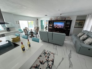 A modern living room with a grey sofa, a dining area with chairs, a TV, and a kitchen area with a coffee machine. - Ultimate Gulf Getaway: Kayaks, Jet Skis and Tours (Cape Coral)
