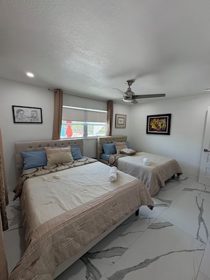 A hotel room with two beds, a ceiling fan, a window with curtains, and artwork on the walls.