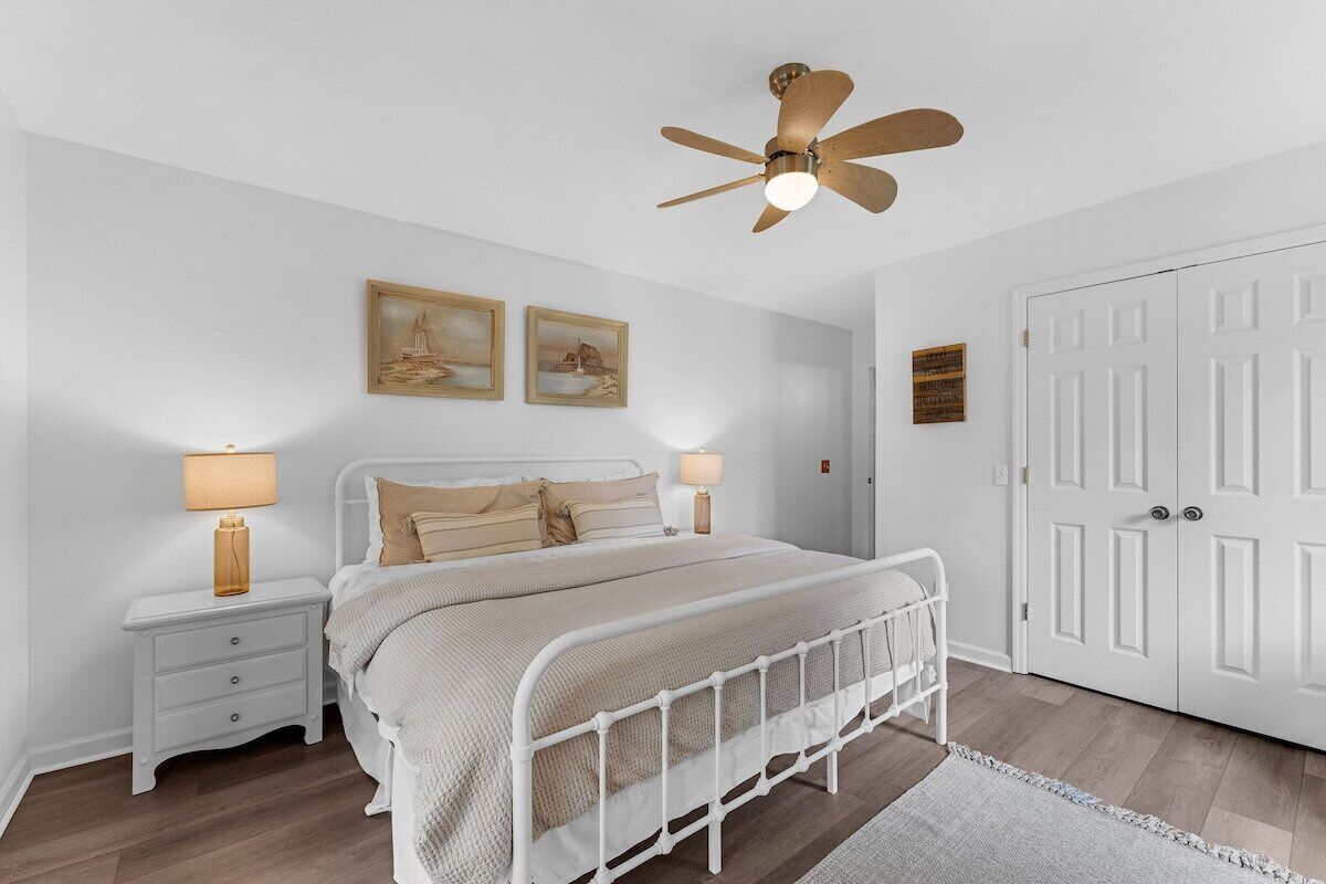 A bedroom with a white metal bed frame, two bedside tables with lamps, a ceiling fan, and a white door.