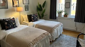 A hotel room with two beds, a large window with curtains, a plant, and framed pictures on the wall.
