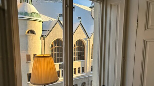 A room with a window overlooking a building with arched windows and a dome. There is a lamp on a table with books and a box.