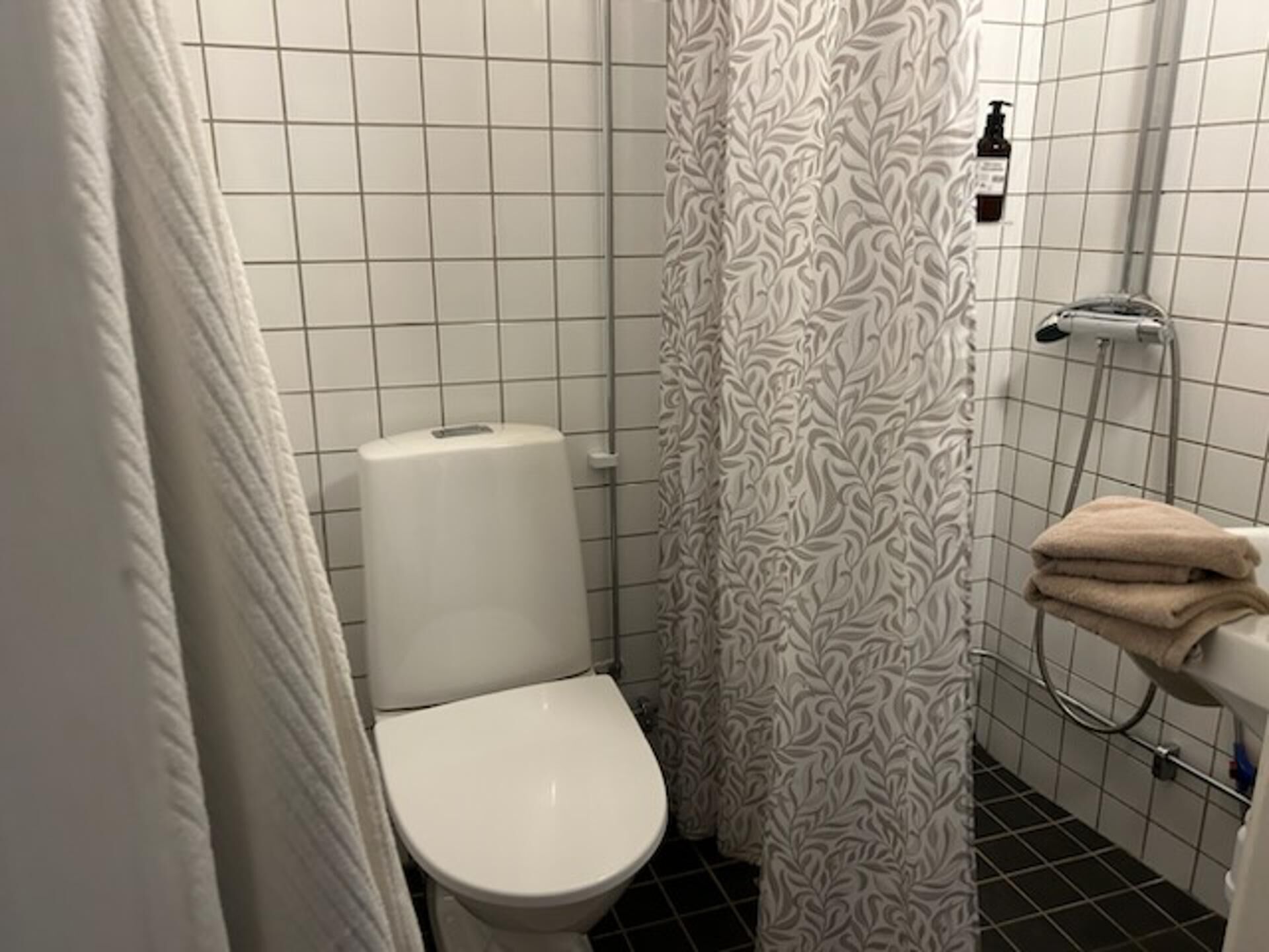 Un baño pequeño con azulejos, un inodoro, cortina de ducha y un perchero con toallas.