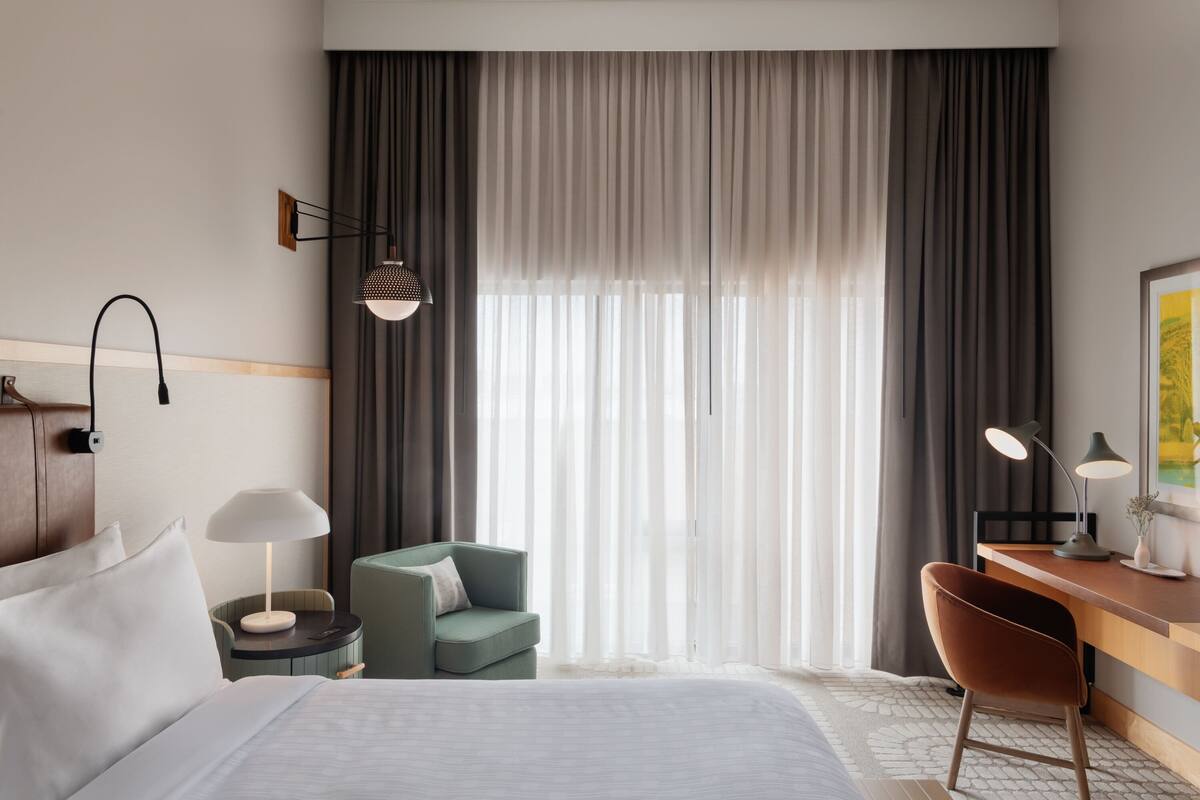 A hotel room with a bed, a desk, a chair, a lamp, and a large window with curtains.