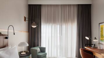 A hotel room with a bed, a desk, a chair, a lamp, and a large window with curtains.