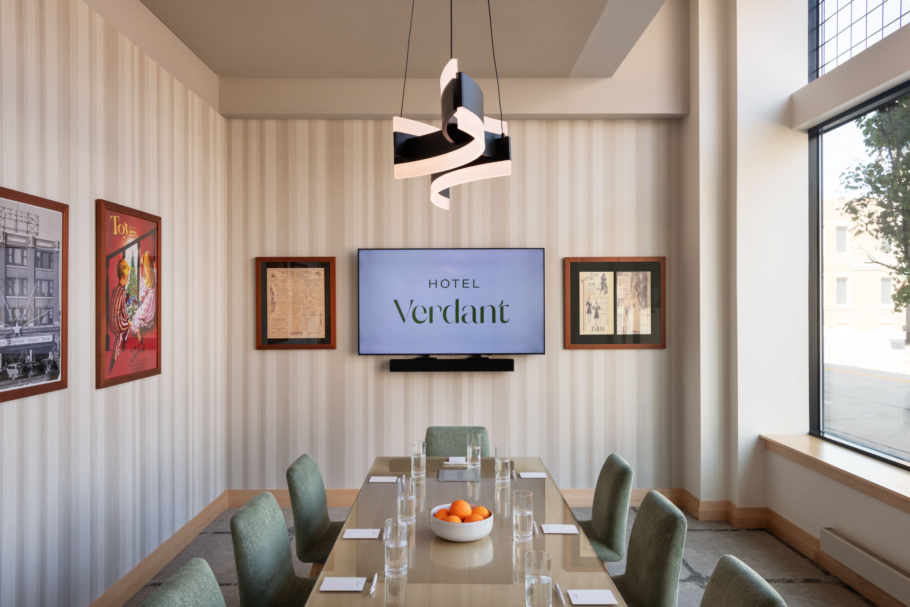 Une chambre d’hôtel moderne dotée d’un grand téléviseur à écran plat affichant le nom « HÔTEL Verdant », d’une longue table à manger rectangulaire avec des chaises, d’un saladier rempli d’oranges et de verres disposés avec soin.