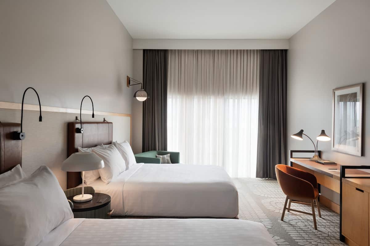 A hotel room with two beds, a desk, a chair, a lamp, and a large window with curtains.