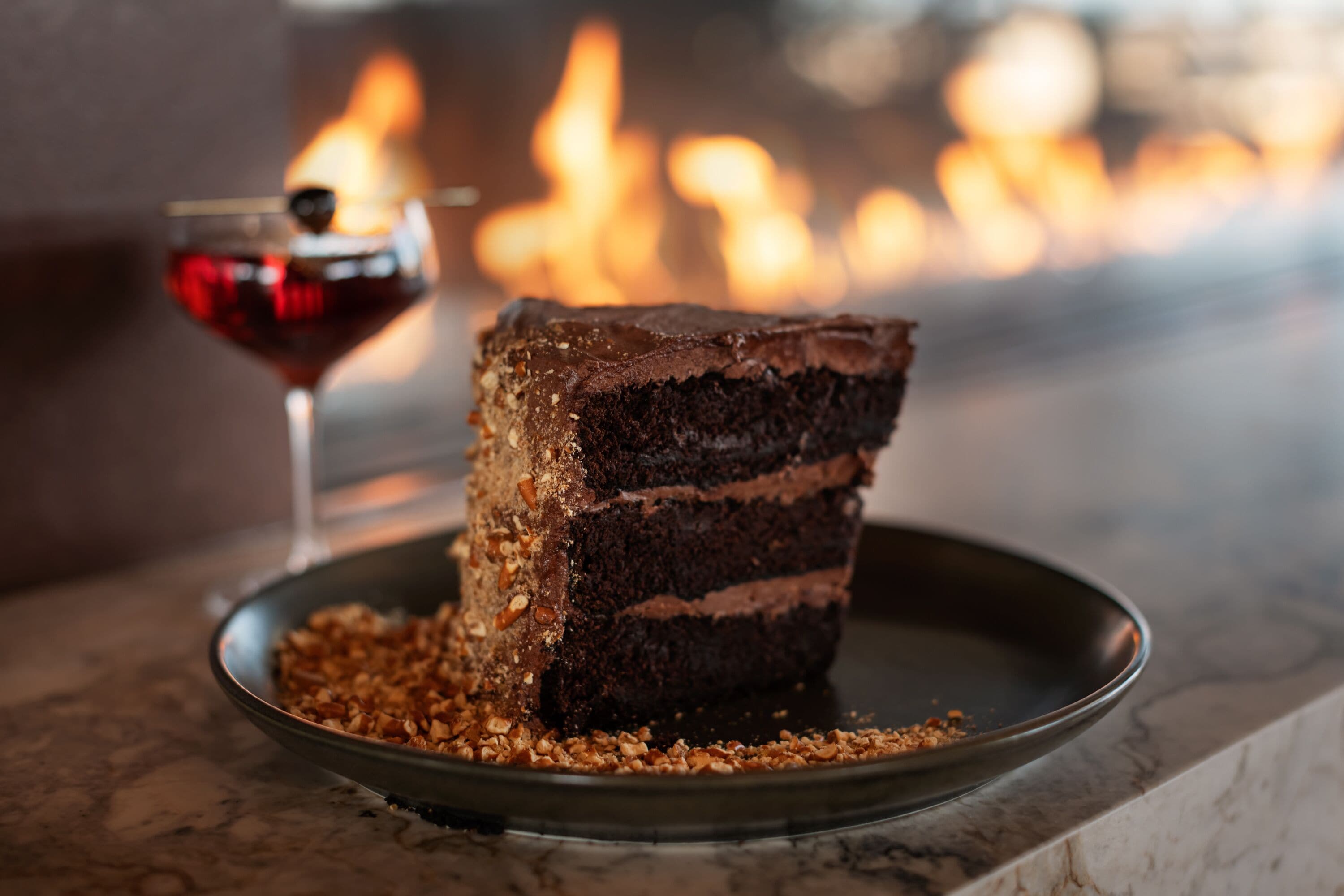 Une part de gâteau au chocolat accompagnée d’un verre de vin rouge, le tout présenté sur une assiette, avec en arrière-plan une image floue d’un feu.