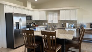 A modern kitchen with white cabinets, a stainless steel refrigerator, a granite countertop, and wooden bar stools. - Beautiful desert house! (Yuma)