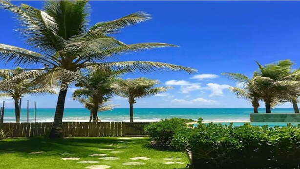 A tropical beach scene with palm trees, a wooden fence, and a clear blue sky.
