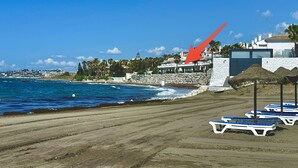 Une plage bordée de rangées de transats, un rivage rocheux et, au fond, un bâtiment coiffé d’un toit en forme de flèche.