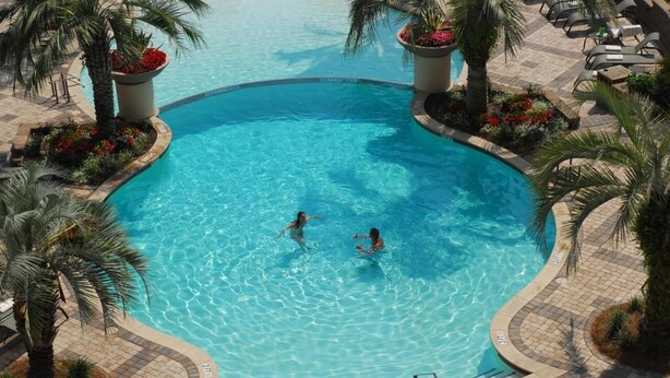 A large, curved swimming pool with two people swimming, surrounded by palm trees and lounge chairs. - Beautiful Waterfront Condo in the Luxurious Four Star Hotel-7th Floor (Myrtle Beach)