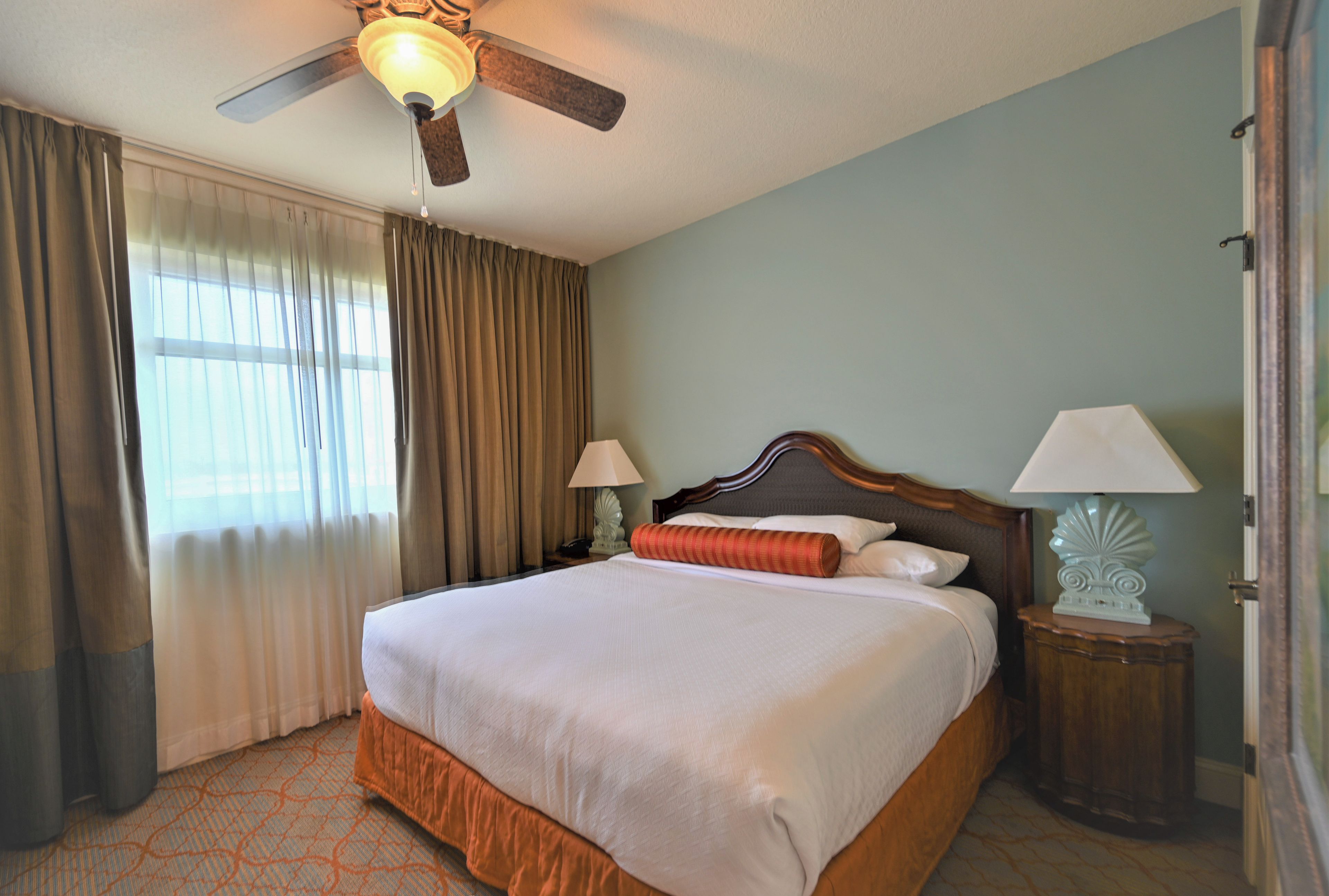 Una habitación de hotel con una cama, dos mesitas de noche, un ventilador de techo y una ventana con cortinas.
