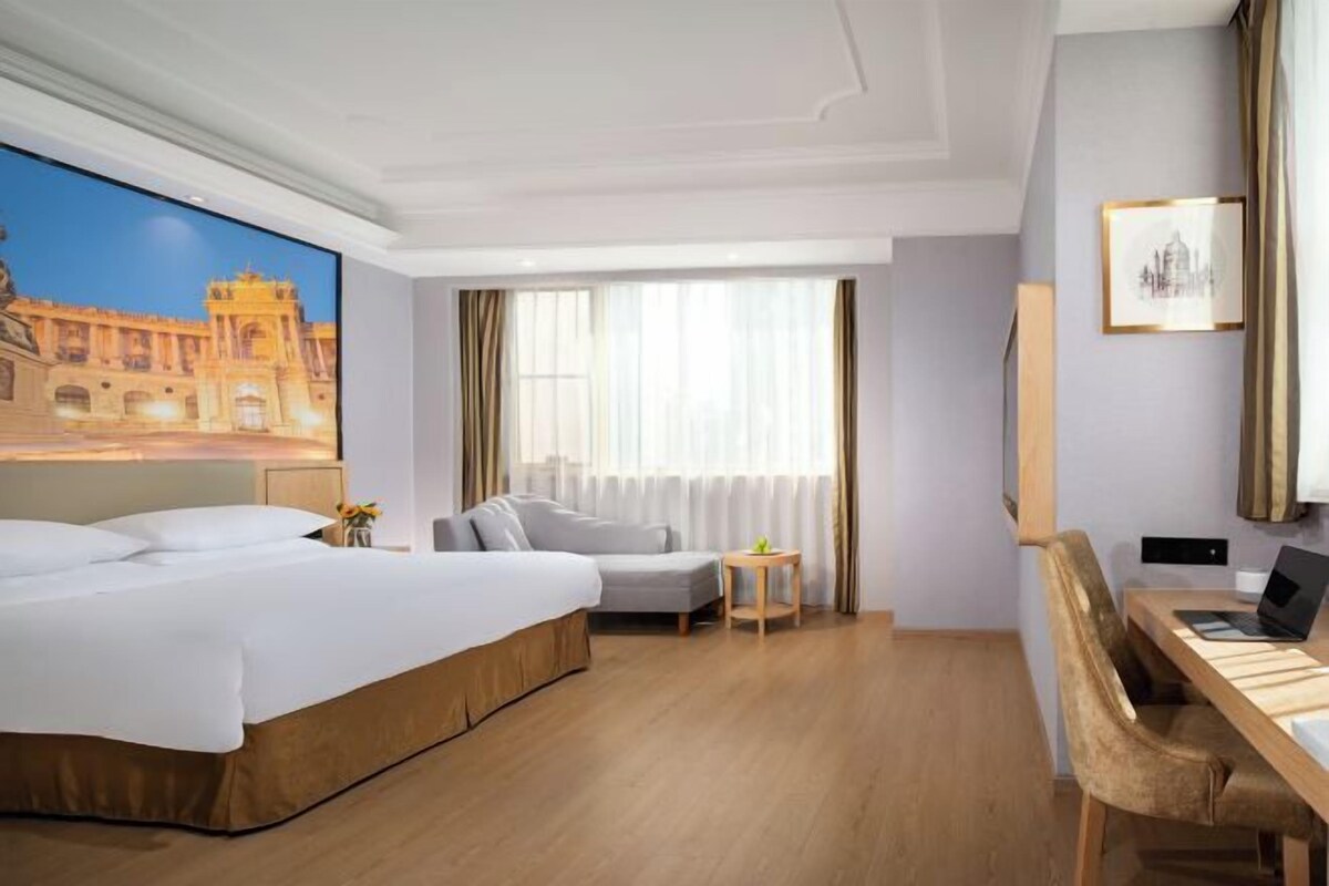 Habitación de hotel con una cama grande, un escritorio, una silla, una mesita y un ventanal con cortinas.