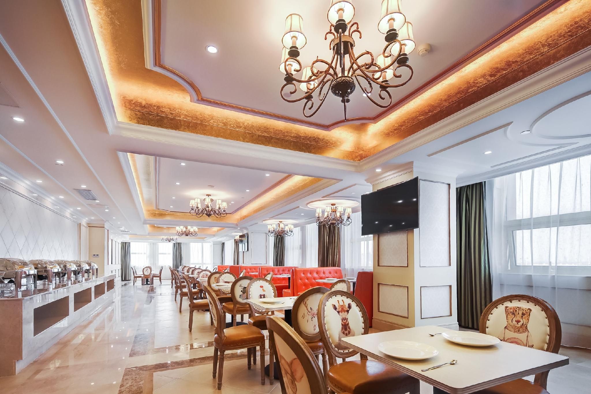A spacious hotel dining area with multiple tables and chairs, a central seating area with red cushioned booths, and ornate chandeliers.