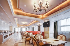 A spacious hotel dining area with multiple tables and chairs, a central seating area with red cushioned booths, and ornate chandeliers.