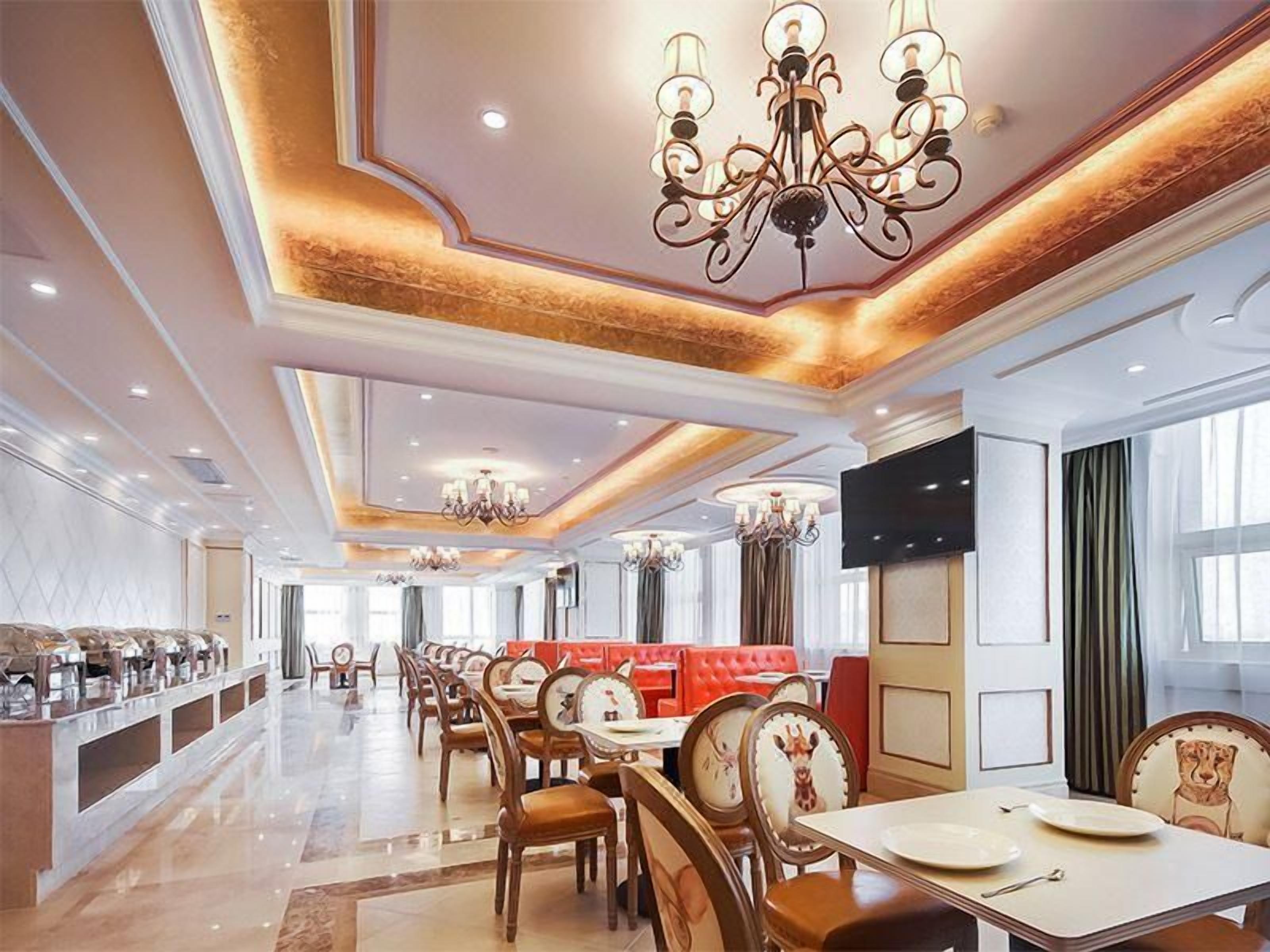 A hotel dining area with multiple tables and chairs, chandeliers, and a television mounted on the wall.