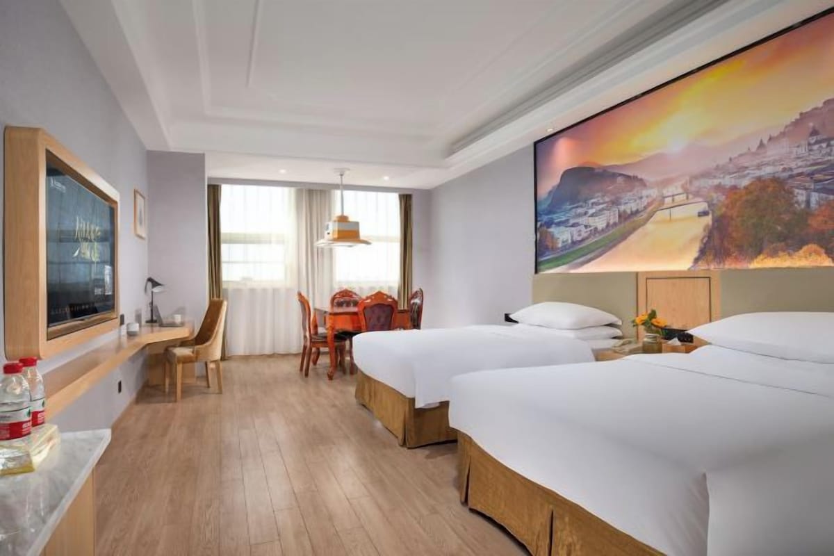 A hotel room with two beds, a dining area, a painting, and a view of a landscape.