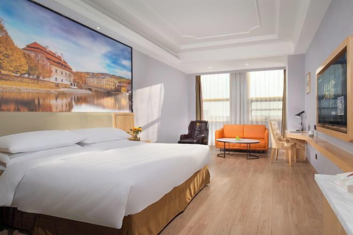 A hotel room with a large bed, a desk, an orange sofa, and a painting of a building by a lake.