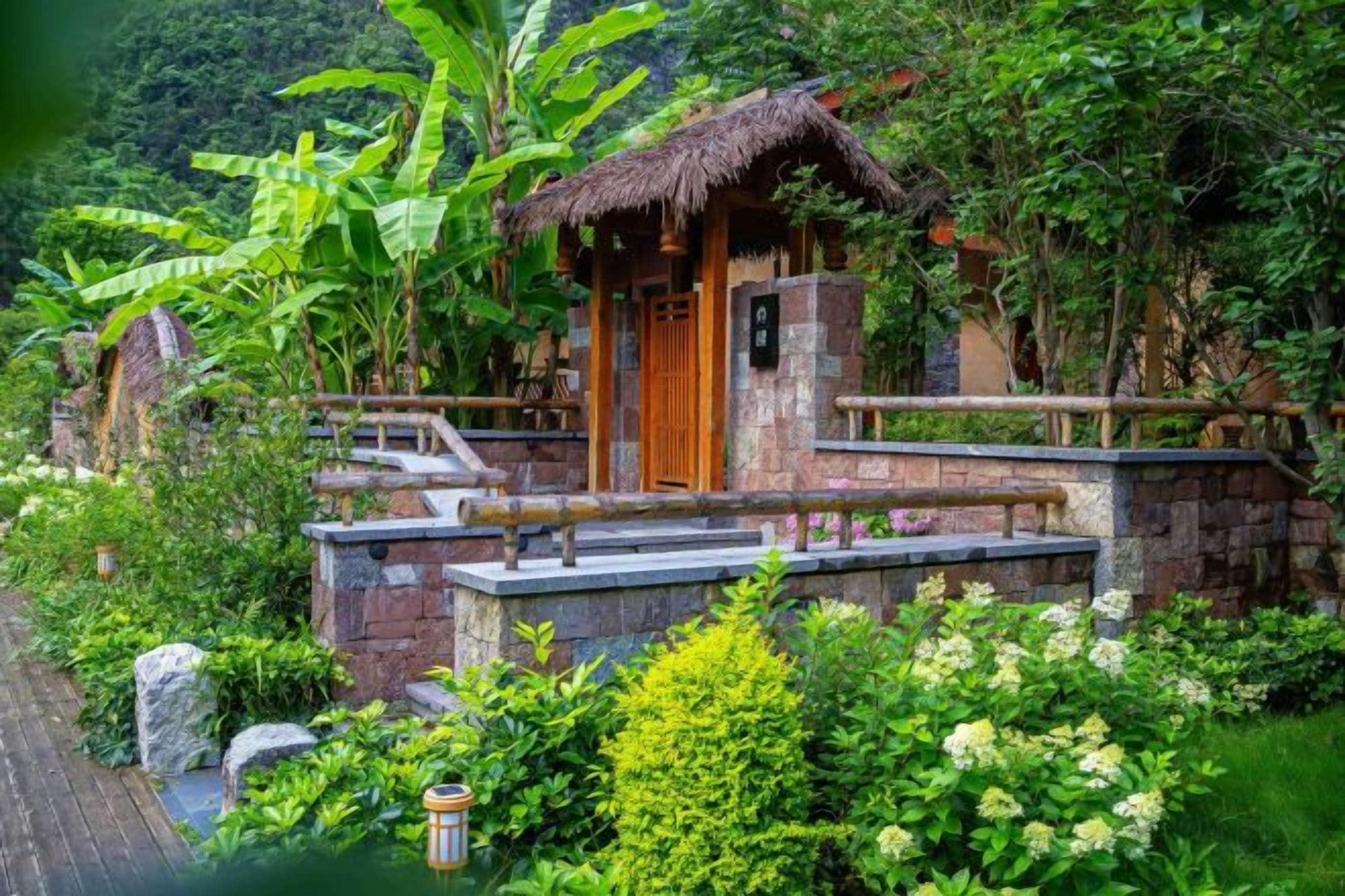 A rustic wooden structure with a thatched roof, surrounded by lush greenery and stone steps leading up to it.
