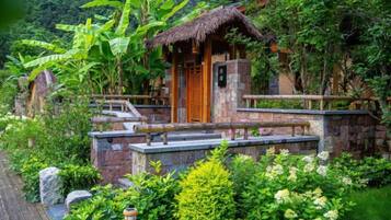 A rustic wooden structure with a thatched roof, surrounded by lush greenery and stone steps leading up to it.