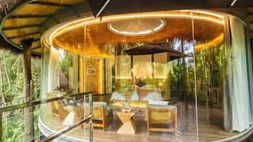 A circular glass-roofed structure with a wooden deck, furnished with chairs and a table, surrounded by lush greenery.