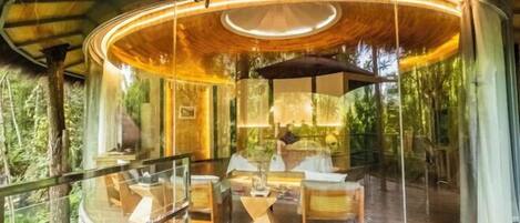 A circular glass-roofed structure with a wooden deck, furnished with chairs and a table, surrounded by lush greenery.