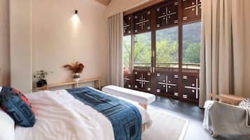 A bedroom with a large bed, a bench, a small table with a vase, and a view of greenery through a window with a wooden screen.