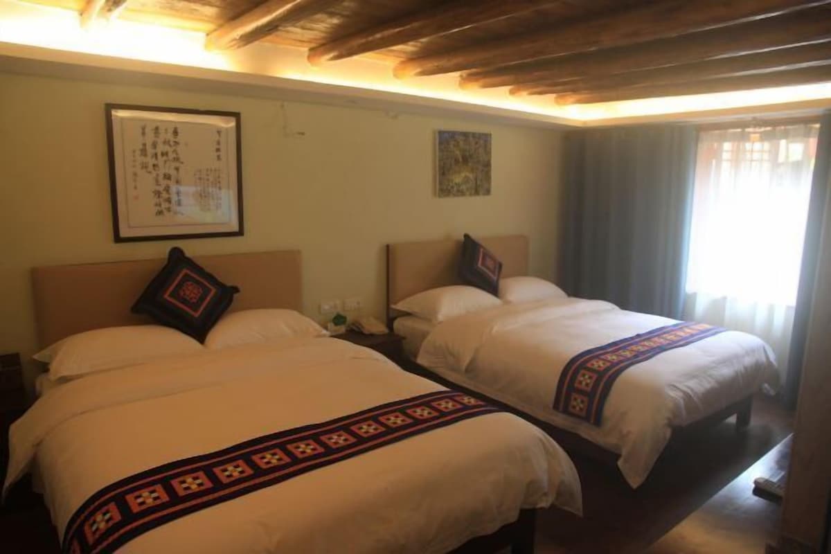 A hotel room with two beds, a wooden ceiling, a framed artwork on the wall, and a window with curtains.