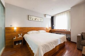 A hotel room with a wooden headboard, a bed with white linens, a bedside table with a lamp, a small desk with a chair, and a window with curtains. - Jinjiang Inn Wuqing Traditional Chinese Medical Hospital Hotel Tianjin (Tianjin)