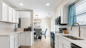 A modern kitchen with white cabinetry, a dining area with a table and chairs, and a large window with blinds. - 1 Block to Beach • Hot Tub • Fire Pit • Game Room (Norfolk)