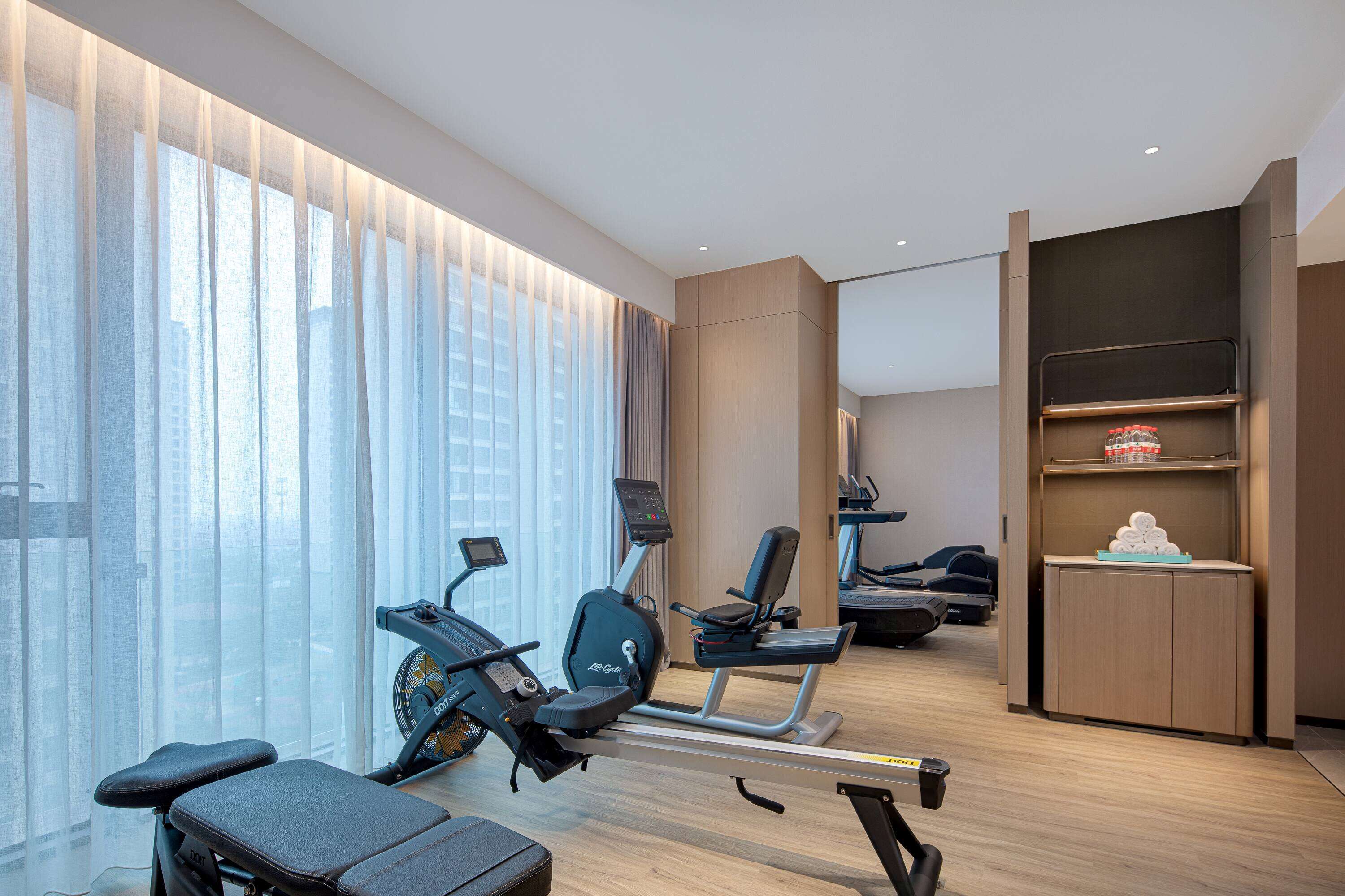 Una habitación de hotel moderna con un ventanal grande, equipo de ejercicio, una kitchenette pequeña y una zona de estar.