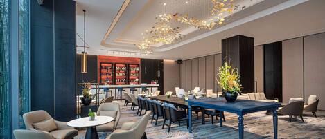A modern hotel lobby with a long dining table, comfortable seating, a bar area, and a decorative ceiling light fixture.