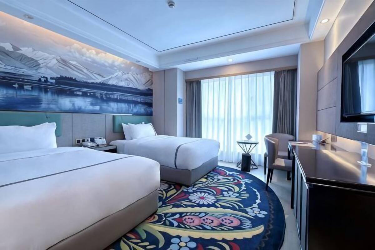 A hotel room with two beds, a desk, a chair, a TV, and a large window with curtains.