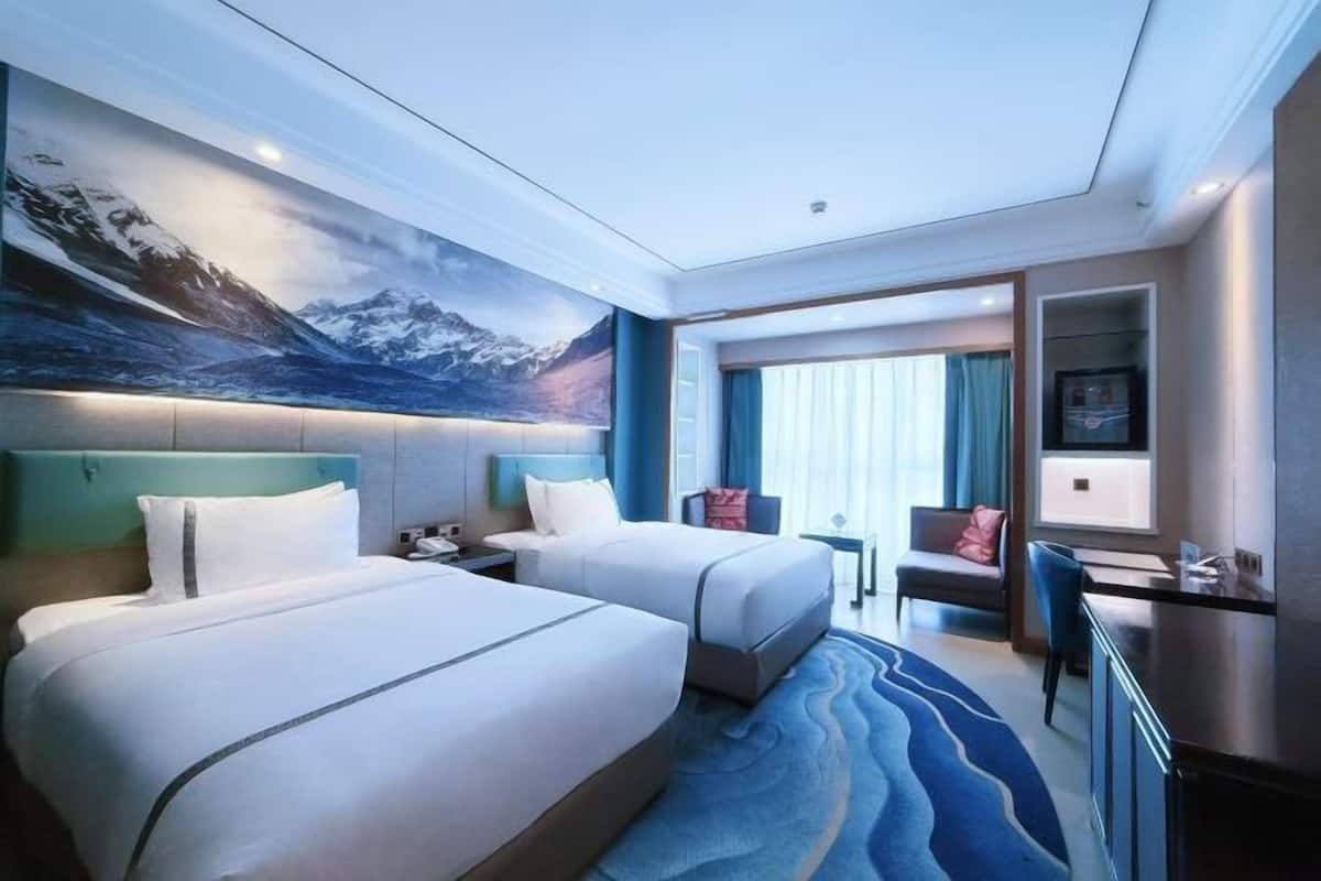 A hotel room with two beds, a desk, a chair, a TV, and a large window with curtains.