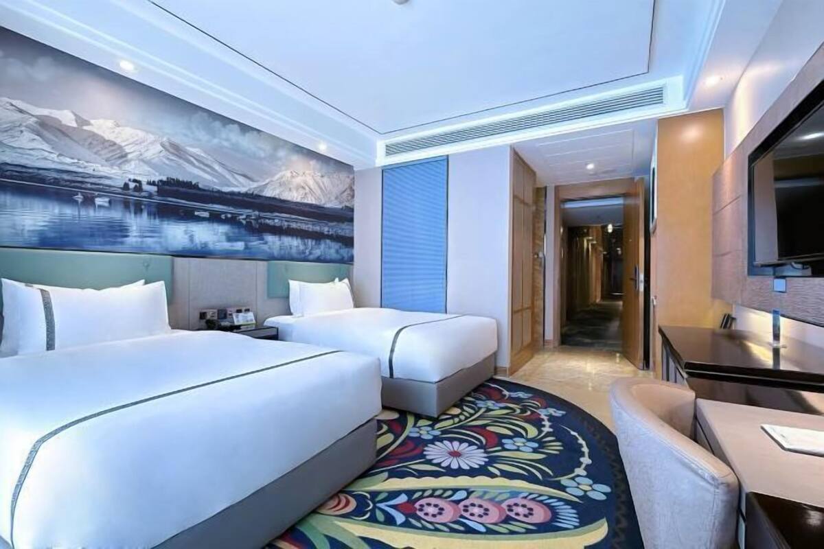 A hotel room with two beds, a large mural of a mountainous landscape, a flat-screen TV, and a patterned carpet.