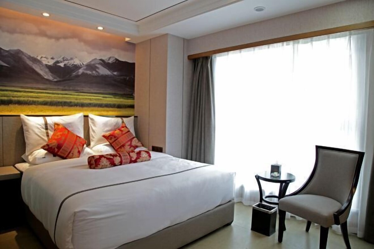 A hotel room with a large bed, a chair, a small table, and a painting of mountains on the wall.