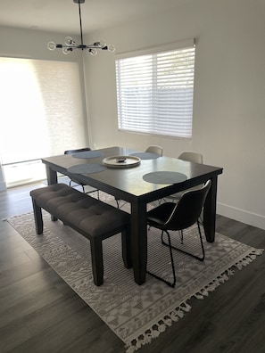 Uma sala de jantar com uma mesa retangular, seis cadeiras, um banco, um candelabro e uma janela com persianas.
