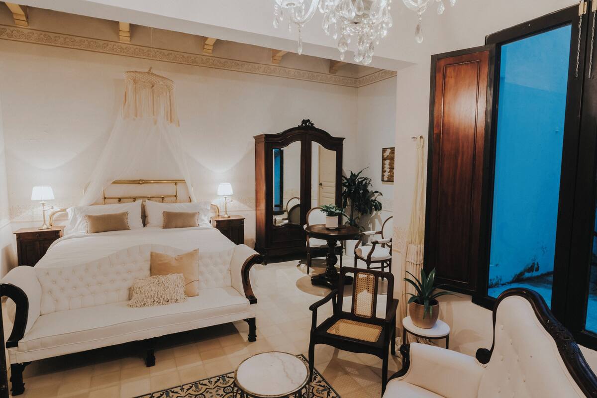 Un dormitorio con una cama grande, una sala de estar con sofá y sillones, una mesita con una planta y un ventanal con vistas.