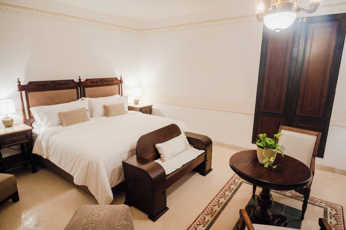 Un dormitorio ordenado con una cama grande, mesitas de noche, un banco de madera, una mesa redonda con una planta y una silla.