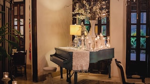 A piano with a white cloth, a table with candles, and a vase with flowers in a room with wooden doors and a chandelier. - Los Murmullos Casa Boutique Campeche (Campeche)