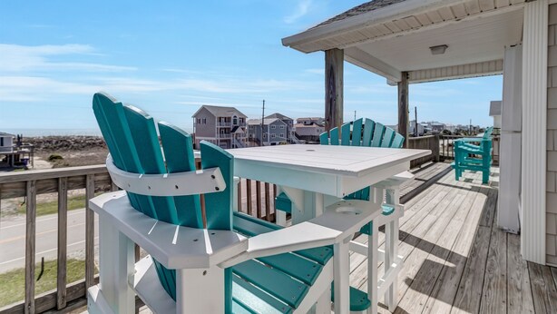 Une terrasse en bois avec une table blanche et des chaises turquoise, donnant sur une plage où l’on aperçoit des maisons au loin.