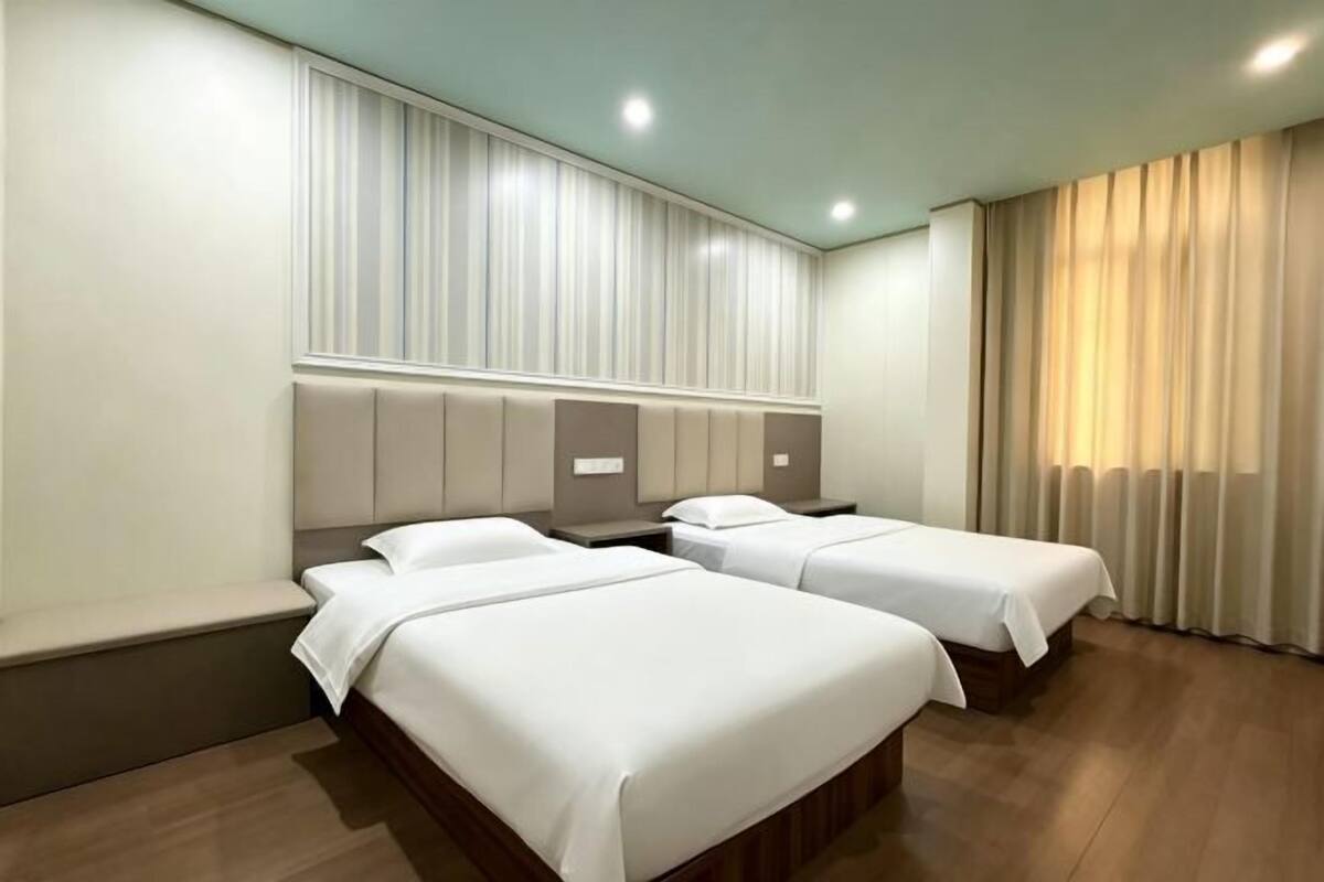 Une chambre d’hôtel avec deux lits, un banc, une tête de lit avec espace de rangement et une grande fenêtre avec des rideaux.