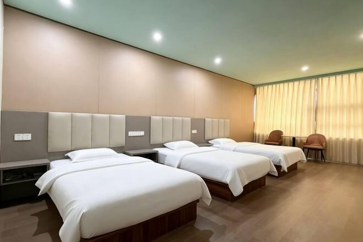 Une chambre d’hôtel avec trois lits, une grande fenêtre avec des rideaux et un coin salon avec des fauteuils.