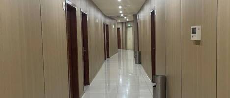 A long, narrow hallway with doors on both sides, a trash bin, and a light switch on the wall.