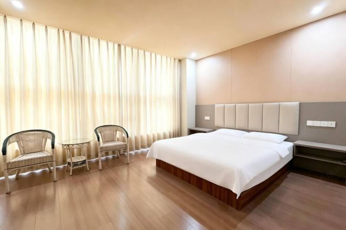 Une chambre d’hôtel avec un grand lit, deux chaises, une petite table et une grande fenêtre avec des rideaux.