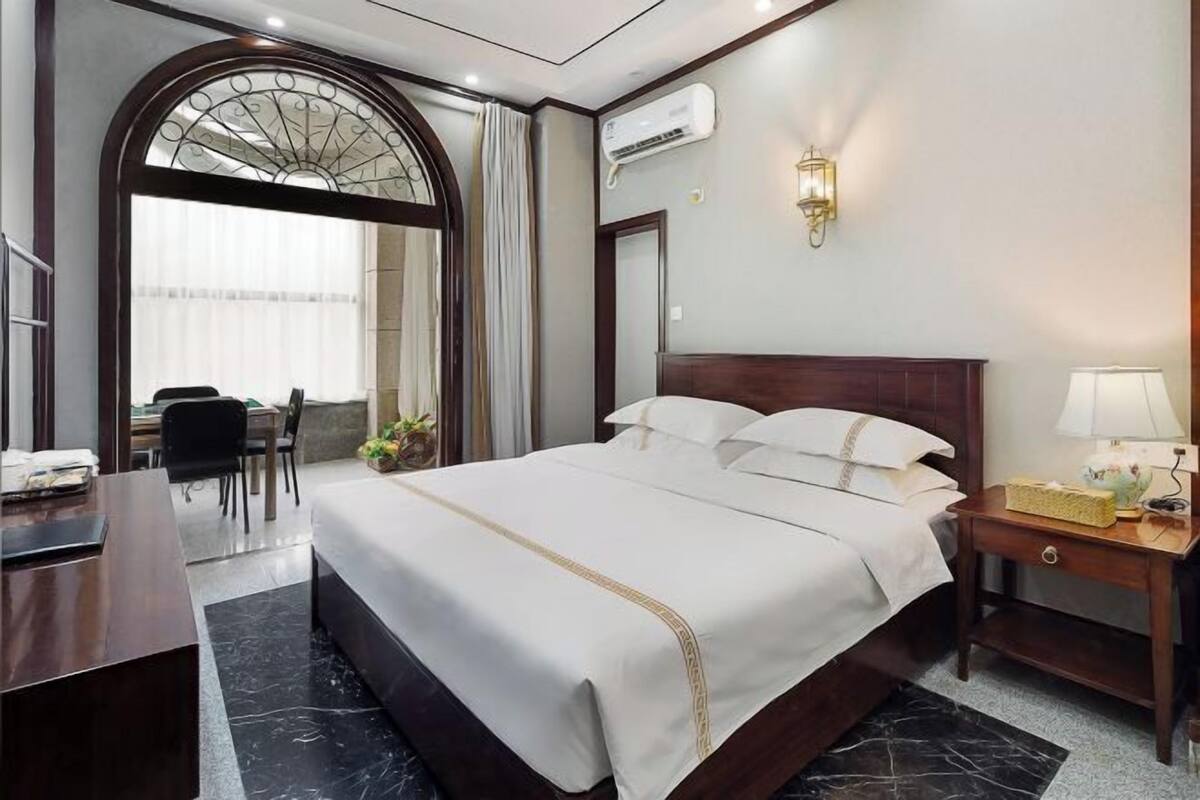 Une chambre d’hôtel avec un grand lit, un bureau, une chaise, une petite table avec une lampe et une fenêtre avec des rideaux.